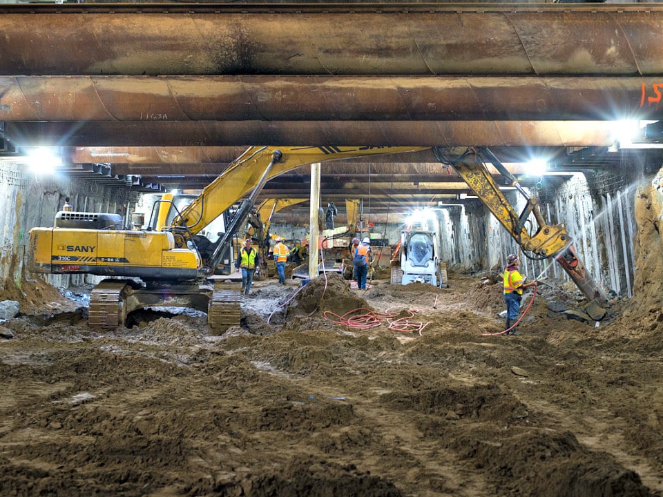 Looking north inside the station box, workers remove excess concrete from the outer walls, exposing steel beams which will be attached to temporary bracing to support the outer walls during excavation.